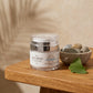 A clear jar of The Alchemy Styrencare SPF 30 Derma-Veil Face Cream sits on a wooden table next to a small bowl of stones and a green leaf, with soft shadow patterns on a beige background.