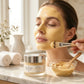 Woman applying gold facial mask with brush, surrounded by skincare products on a marble surface.