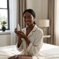 Woman in a white robe holding a skincare bottle in a bedroom setting