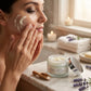 Woman applying cream to her face with a jar of cream on a marble surface.