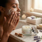 Woman applying cream to her face with a jar of cream and lavender flowers on a marble surface.