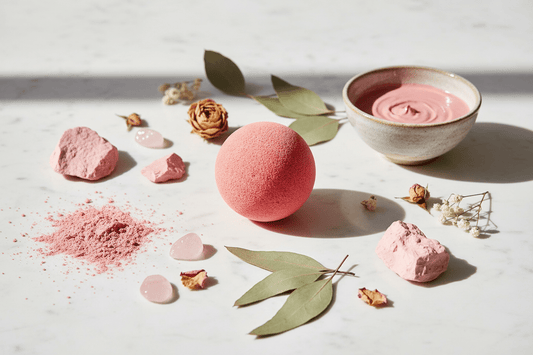 A pink bath bomb sits on a white marble surface surrounded by decorative leaves, dried flowers, and small pink stone pieces, with a small bowl of matching pink cream in the background.