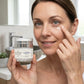 A woman in a bathroom holds a jar of skincare cream while applying a dollop of the cream to her cheek. The jar is labeled 'THE ALCHEMY OF AGE' and 'Eye Cream'.
