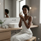 Woman in a bathroom holding a skincare product in front of a mirror.