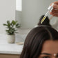 Person applying a dropper of oil to their hair, with a blurred indoor background.