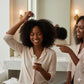 Two women in a bathroom setting, one applying hair product and the other pointing at it.