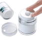 A clear cylindrical cosmetic jar with a white screw-top lid and a small white central button, shown with a hand pressing that button. A separate clear lid and another jar are visible on a plain white background.