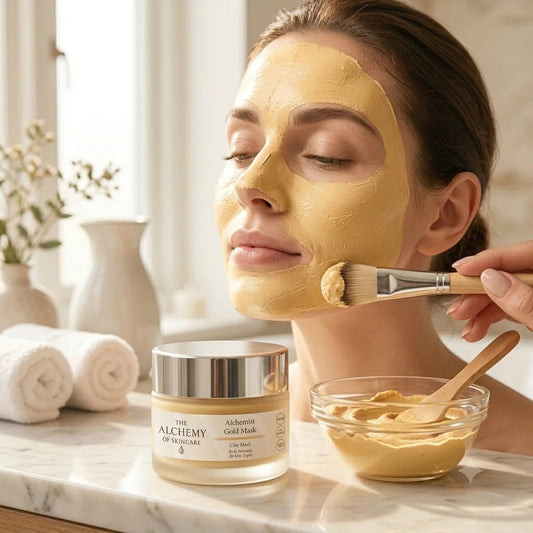 Woman applying gold facial mask with brush, surrounded by skincare products on a marble surface.