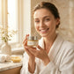 Woman in a bathrobe holding a skincare product in a cozy bathroom setting